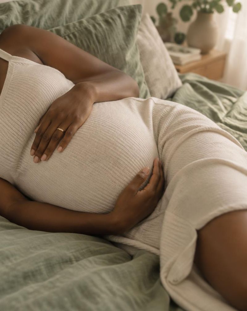 Pregnant woman resting on her side with her hands on her bump, on soft sage bedding
