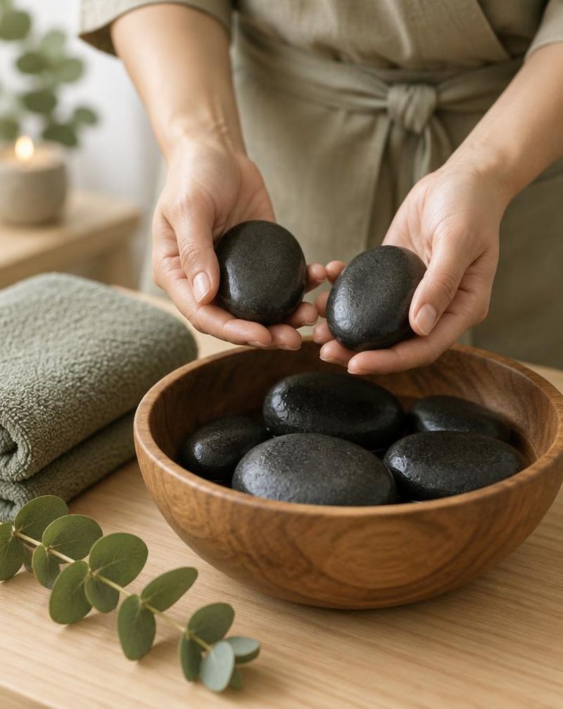 Therapist's hands holding warm basalt stones over a wooden bowl, with a sage towel and eucalyptus